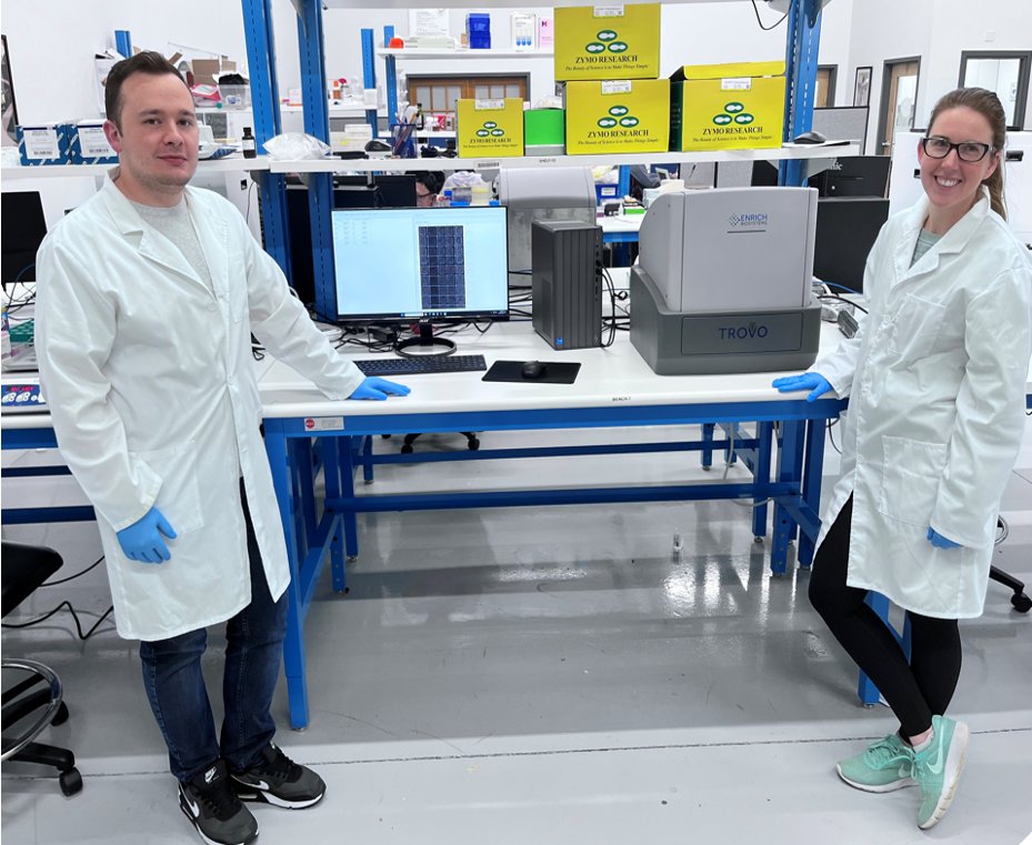 Two individuals wearing white lab coats and blue gloves stand next to a workstation in a laboratory. The table holds scientific equipment, including a device labeled TROVO and a monitor displaying analytical data. The laboratory features a clean and organized layout with shelves and research materials in the background.